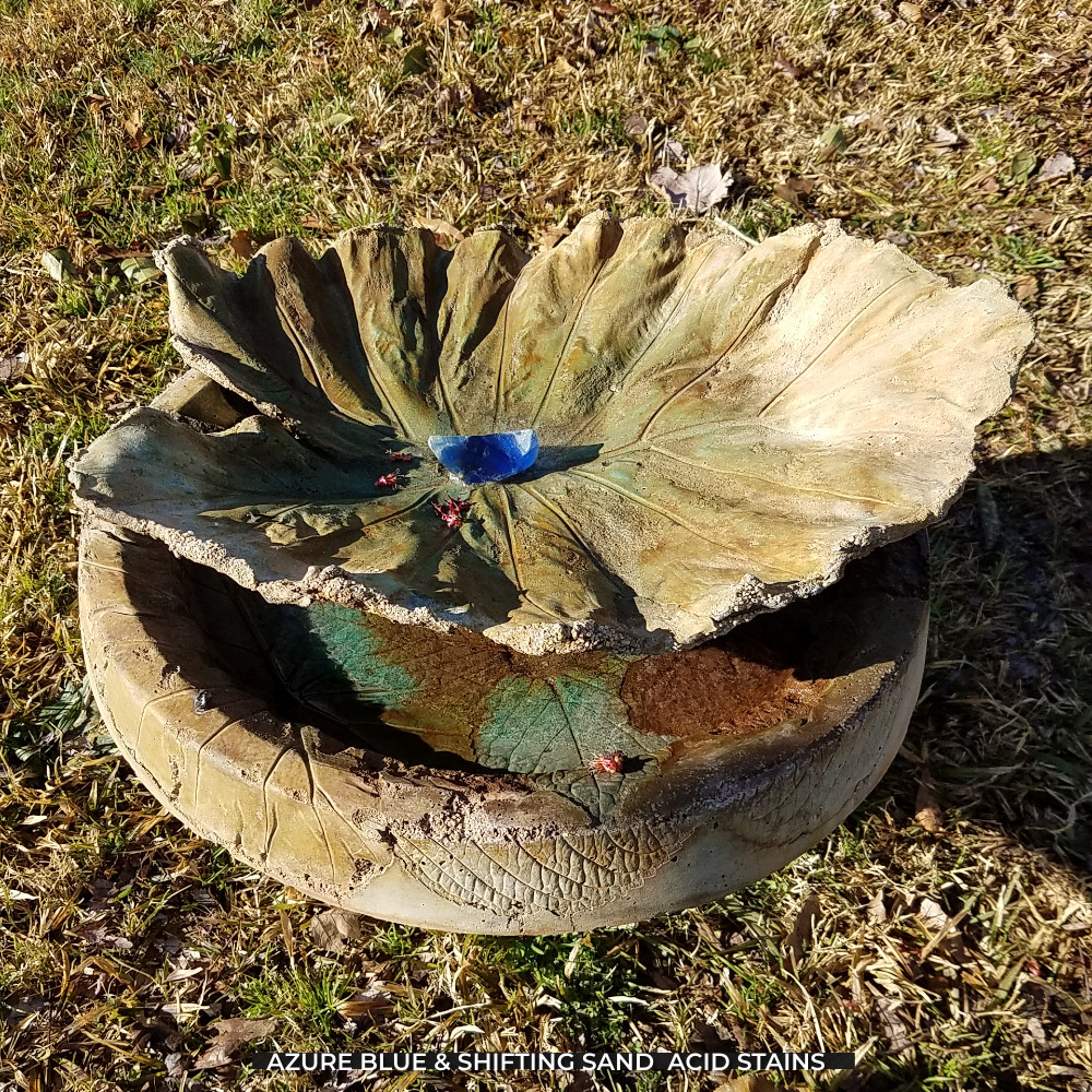 A picturesque bird bath stained with Shifting Sand acid stains for the base and Azure Blue accents highlighting its artistic features. The bird bath is structured like a bowl, with a large, intricately designed concrete leaf mounted on top, providing an inviting spot for the birds.