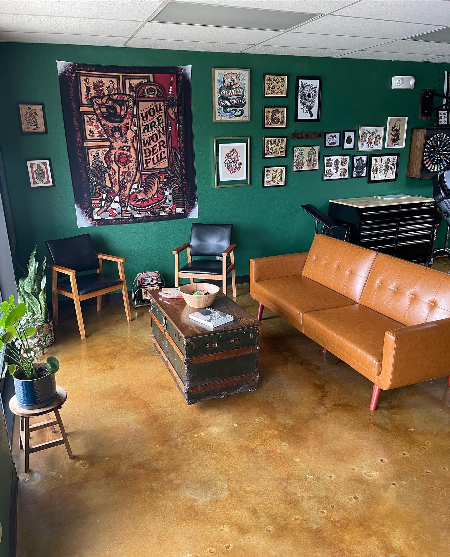 A tattoo studio waiting area with a polished concrete floor in warm amber and brown tones, surrounded by vintage decor, leather seating, and artwork on the walls.