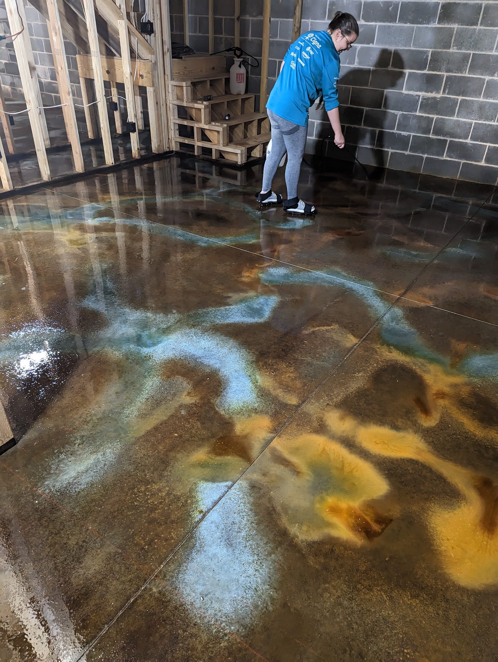 Woman applying three different acid stains - Coffee Brown, Malayan Buff, and Seagrass - onto the concrete basement floor using a pump sprayer