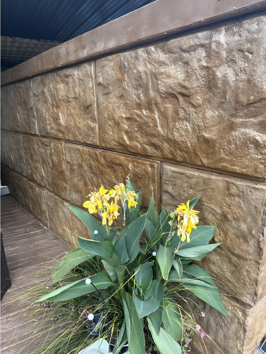 Stamped concrete retaining wall finished with Café Royale EasyTint™ tinted sealer, showing deep golden-brown tones and a natural stone texture beside green plants